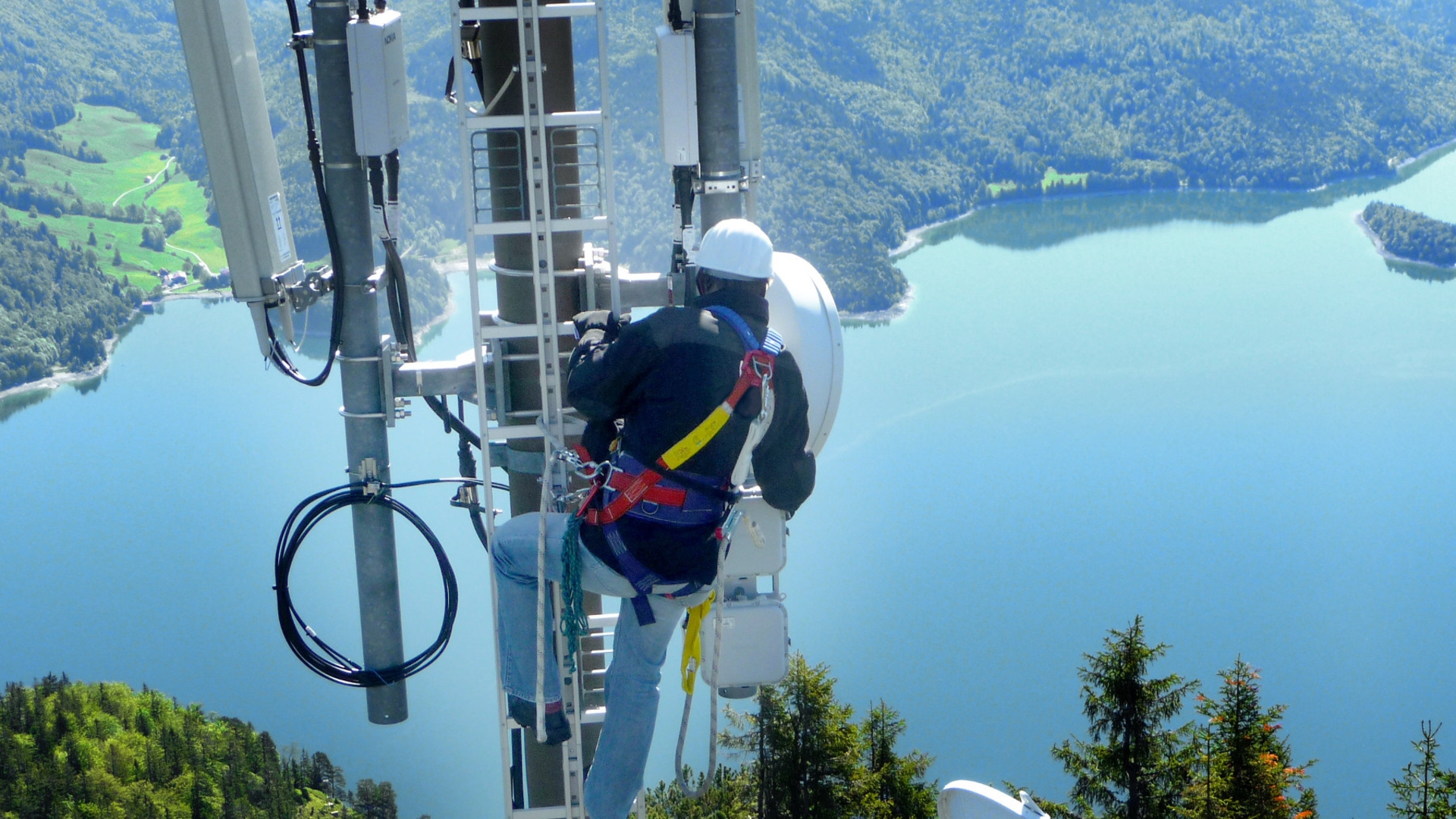 Techniker arbeitet in großer Höhe an Funkmasten vor Bergsee-Panorama