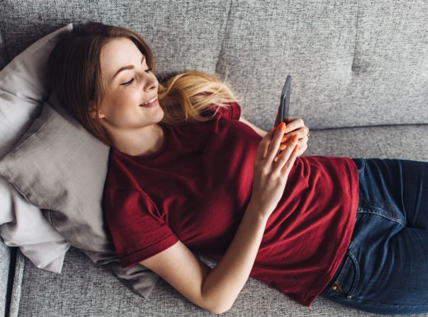 Frau tippt lächelnd auf der Couch auf ihr Handy