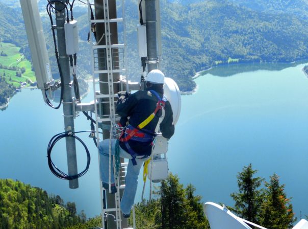 Techniker arbeitet in großer Höhe an Funkmasten vor Bergsee-Panorama