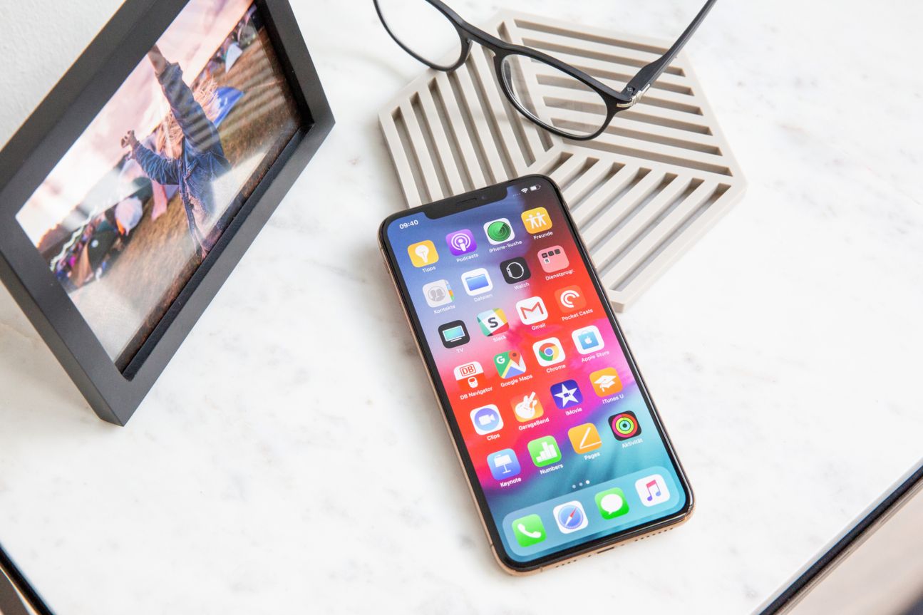 Das iPhone Xs Max auf einem Schreibtisch neben einem Bilderrahmen und einer Brille.
